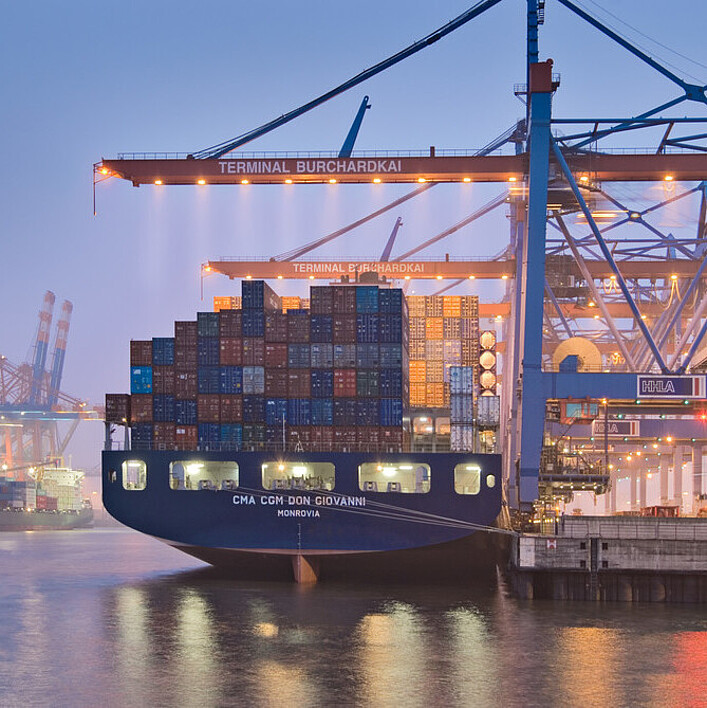 Containerschiff am Terminal Burchardkai im Hamburger Hafen bei Abenddämmerung, umgeben von großen Containerkränen.
